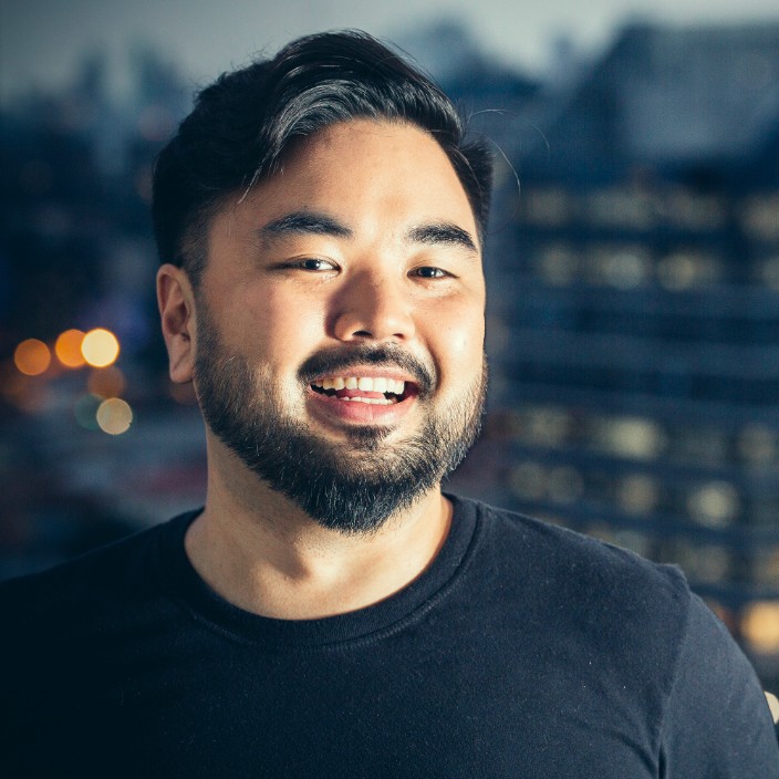 Homem adulto de ascendência japonesa, com cabelo curto e barba aparada, olhando levemente para a lateral com expressão natural e sorriso aberto. Veste camiseta preta, em um enquadramento próximo (retrato), com foco no rosto. Ao fundo, uma paisagem urbana desfocada com luzes difusas, sugerindo fim de tarde ou início da noite. A iluminação é suave e lateral, criando contraste leve e destacando os traços do rosto, transmitindo proximidade sem perder precisão.